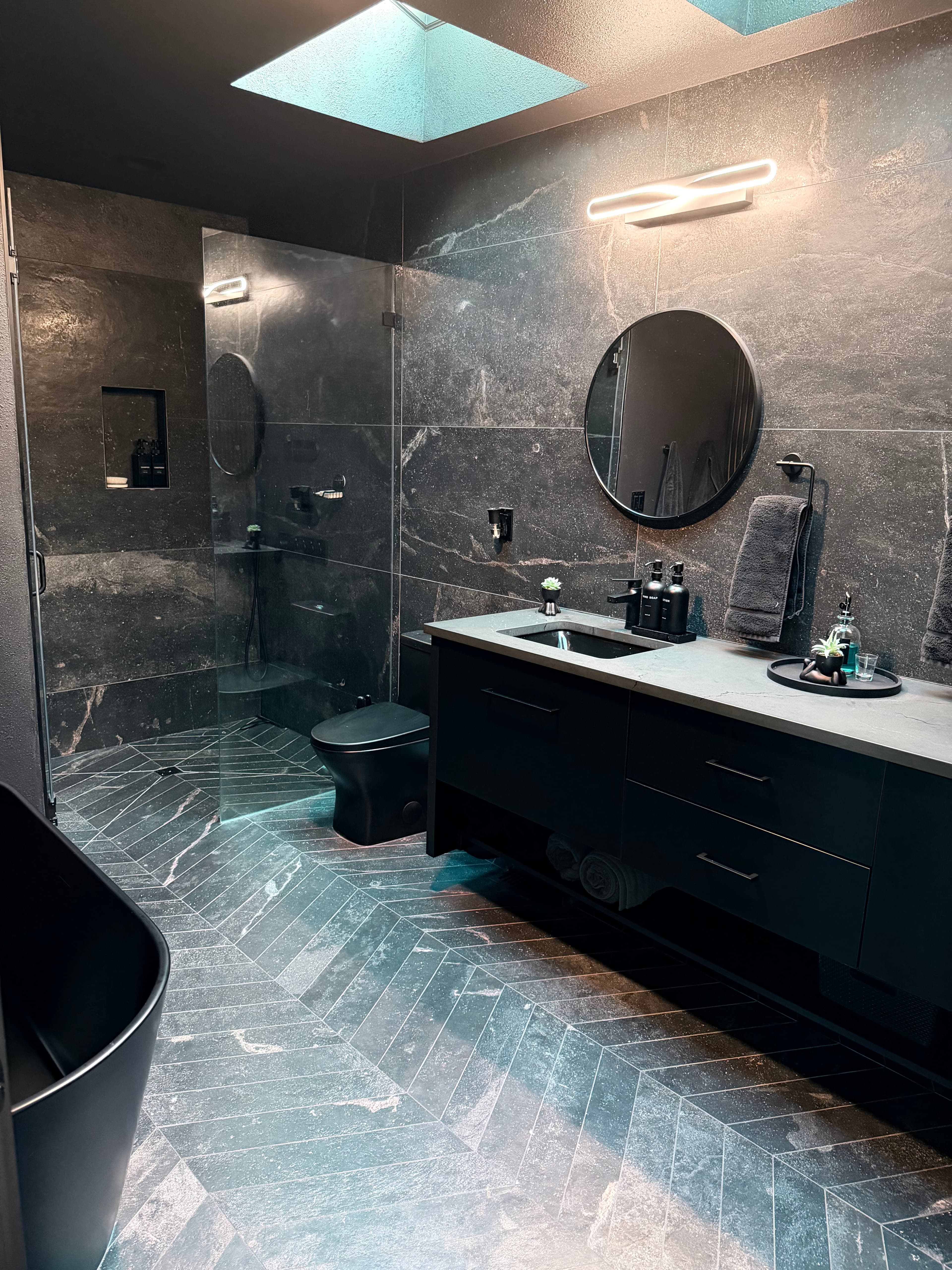Bath Project Photo — Bathroom remodel in dark marble-look tile with a round mirror, black fixtures, and a skylight
