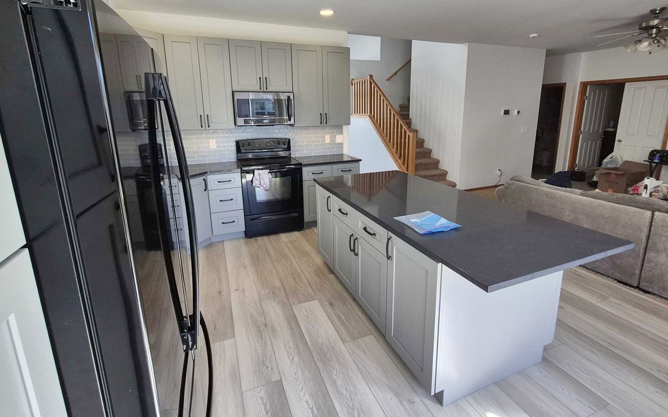 Gray kitchen with island, cabinets, and appliances. Light wood floor, stairs in background. Gray kitchen with island, cabinets, and appliances. Light wood floor, stairs in background.
